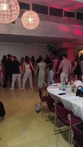 A group of people are holding hands and dancing in a circle. There are two large spherical light fixtures hanging from the ceiling. In the foreground, there is a round table covered with a white tablecloth, with chairs around it. On the table, there are bottles, glasses, and some decorative items. The room appears to be dimly lit with pink and red lighting.