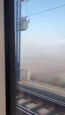 The video shows the view from inside a moving train through a window. The outside environment is shrouded in thick fog or dust, with visibility significantly reduced. The landscape consists of green fields and sparse vegetation, partially obscured by the haze. Overhead power lines are visible against the pale blue sky. The train tracks run parallel to the window, and the train appears to be traveling at a moderate speed.
