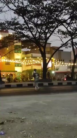 A low-angle shot captures an outdoor scene at dusk, illuminated by strings of warm lights decorating buildings. A person is walking away from the camera on a sidewalk, dressed in a dark t-shirt and light-colored pants. A motorcycle speeds by on the road in the mid-ground, its motion blurred. Further down the road, a car with its headlights on is visible, also in motion. Trees with dark, bare branches extend over the scene from the top of the frame. The overall impression is of a street at twilight, possibly a commercial area, with ongoing activity.