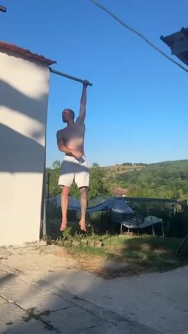 A young, muscular man with tattoos on his leg is performing a static hold on a horizontal bar. He is hanging from the bar with one arm fully extended, while his body is angled sideways. He is wearing white shorts. The scene is set outdoors on a sunny day with a clear blue sky. Behind the man, a white building with a red tiled roof is visible, along with some greenery and rolling hills in the distance. There are also some chairs covered with striped tarps in the background, suggesting a backyard or outdoor seating area. The overall impression is one of a casual, active moment during a warm day, possibly in a rural or semi-rural location given the natural landscape. There is no visible text in the media file.