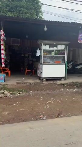 The video pans across a street scene. The first few seconds show a storefront with a yellow sign reading "CISEWAXAN" and phone numbers. A dark tarp is visible on the left. The next segment shows a closed storefront with wooden shutters and a motorcycle parked in front. A juice stand with colorful fruits and prices is then displayed, followed by a building with an awning and plants. The final shot shows a residential building with a black car parked in the garage. The environment appears to be an urban or semi-urban area with a mix of commercial and residential properties. The street is unpaved in some sections, with grass and debris. The sky is overcast. There are no people visible in the video.