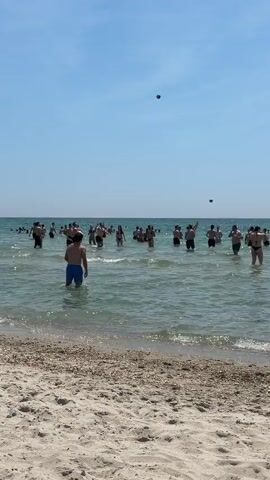 A wide shot captures a crowded beach scene on a clear, sunny day. In the foreground, light brown sand transitions into clear, light blue-green ocean water. Numerous people, predominantly young adults, are standing in the ocean, with the water level reaching their waists to chests. They are actively engaged in playing a game with a dark-colored beach ball, which is frequently seen in the air. Individuals are observed moving, reaching, and splashing as they interact with the ball. One person in blue swim trunks stands closer to the shore, facing the larger group in the water. The sky above is bright and cloudless.