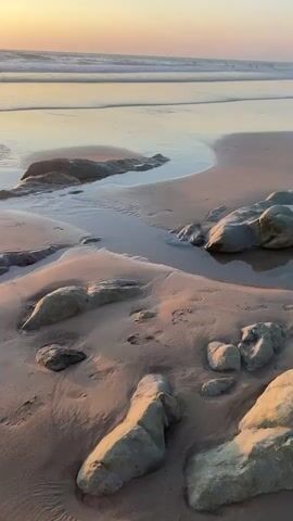 The video shows a beach scene at sunset. The camera is positioned low to the ground, focusing on the sand and some rocks. In the distance, a group of people are standing in the shallow water near the shore, with waves gently breaking. The sun is low on the horizon, casting a warm, golden light across the sky and reflecting on the wet sand. Footprints are visible in the sand.