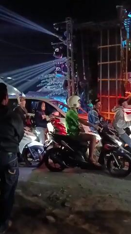 The video shows an outdoor event at night. In the foreground, a person stands on the left side, facing away from the camera. Several motorcycles are lined up in the middle ground. On the motorcycles are various people, some wearing helmets, and some are looking towards the right. Behind the motorcycles, a white vehicle is visible. To the right of the vehicle, a large speaker system on a metal truss is illuminated with red lights, and bright white and blue lights are projected into the night sky from the left side of the frame. A few people are walking around in front of the speaker system. The ground appears to be dirt or gravel.
