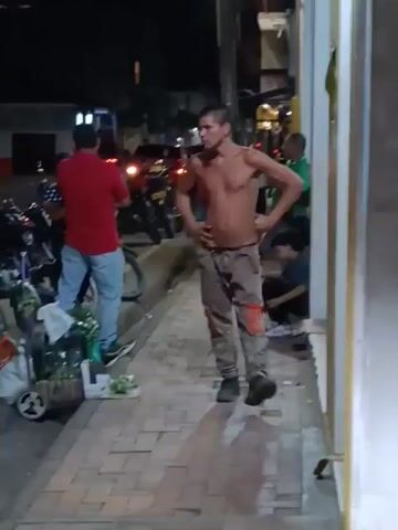 The video shows a street scene at night. A shirtless man in camouflage pants and boots is standing on the sidewalk with his hands on his hips. To his left, a man in a red shirt and jeans is looking at a parked bicycle and a cart with various items on it. In the background, there are parked motorcycles, cars with their lights on, and other people. The sidewalk is tiled. The lighting is provided by streetlights.