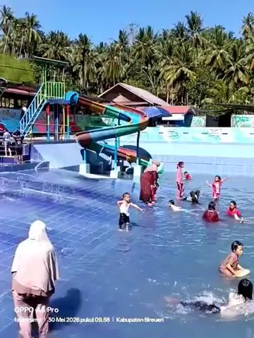 A low-angle shot shows a group of children and adults in a shallow swimming pool. A blue, green, red, and yellow waterslide stands on the left side of the frame, next to a staircase. In the center of the pool is a mushroom-shaped fountain with white spots on a red top. The pool is lined with blue tiles, and the water is light blue. Children are standing, swimming, and playing in the water. Some are wearing swimsuits, while others are wearing casual clothing. Adults in modest clothing are present, some near the children. The background features lush greenery, including numerous palm trees, and some buildings can be seen through the foliage. The sky is bright blue, indicating a sunny day. At the bottom of the frame, text displays "OPPO A6 Pro fataelrumy - 30 Mei 2026 pukul 09.58 - Kabupaten Bireuen."