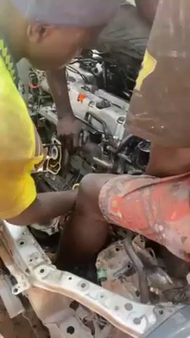 Three men are working on an engine in the front of a car. The car frame is white and the engine is silver and black. The men are standing around the engine bay, reaching in to work on the engine. The man on the left is wearing a yellow shirt and a light grey cap. The man on the right is wearing patterned shorts and a dirty shirt with yellow writing. The third man is mostly hidden behind the engine. The scene appears to be outdoors.