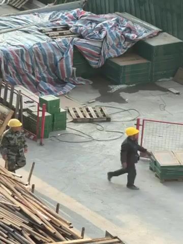 The video shows an outdoor construction site. Two workers wearing yellow hard hats are visible. One is walking across the concrete surface carrying something, while the other is standing near a stack of wooden planks. A large stack of materials covered with a striped tarp sits to the left of the frame. There are also stacks of wooden pallets, green boxes, and red barriers around the site. The ground appears to be wet.