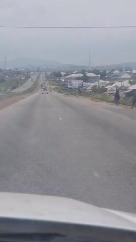 The video shows a road in a suburban or rural area. It's a cloudy day, and the road appears relatively empty, with only a few cars and a single motorcycle visible. On the right side of the road, there are some houses and a billboard that is visible. In the distance, there are hills and transmission towers. The camera is positioned inside a moving vehicle, looking out through the windshield.