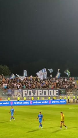 This is a vertical, low-angle video of a nighttime soccer match. The field is green, and in the background, a section of stadium seating is filled with cheering fans waving flags and holding banners. The banners read "BOJDONE" and "OPŠTINA NIKŠIĆ." Three soccer players in blue and yellow uniforms are visible on the field. The sky above is dark.
