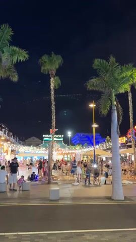 The video shows a lively nighttime scene in what appears to be a resort or entertainment area. In the foreground, there are two large palm trees on either side, their trunks wrapped in fairy lights. The ground is paved, with a road visible in the very bottom. In the middle ground, a pedestrian area is bustling with activity. People are walking, standing, and sitting at tables outside what seems to be a restaurant or bar. Some tables are covered by umbrellas. A sign for "MINI GOLF" is visible on a building in the background, also decorated with lights. Further in the background, a building is illuminated with bright blue lights. The overall atmosphere is festive and vibrant, with numerous lights and the presence of many people enjoying the evening. The sky is dark, indicating it is nighttime, and there are faint lights or possibly birds visible high in the sky. The video is shot from a slightly low angle, looking up towards the scene.