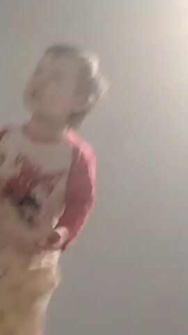 A young child in pajamas stands on a bed covered with a patterned comforter. The child jumps up and down a few times before briefly sitting. The camera is positioned low, looking up at the child. The background shows a plain wall, a corner of curtains, and a bedside lamp. The lighting is soft and somewhat diffuse.