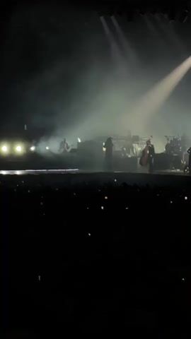 The video shows a live music performance on a stage, with a band playing and a lead singer standing center stage. The stage is illuminated by bright spotlights, with fog or smoke creating a hazy atmosphere. The band appears to consist of a guitarist, a keyboard player, a double bass player, and a drummer. The audience is visible in the foreground, holding up their phones to record the performance.