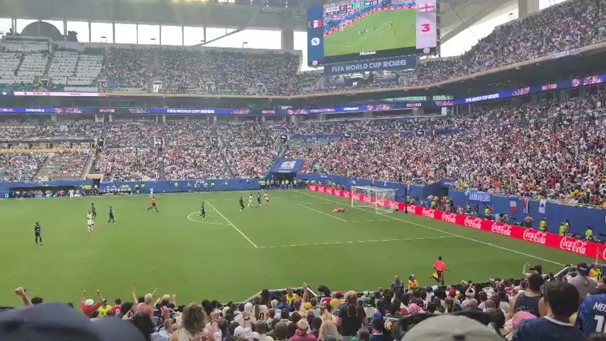 The video shows a wide shot of a soccer stadium filled with spectators. In the foreground, the arms and hands of several fans are raised in celebration. They appear to be cheering and excited. In the background, the soccer field is visible with players on it. A large screen at the top of the stadium displays the score, with France leading England 3-1. The overall atmosphere suggests an intense and exciting soccer match.