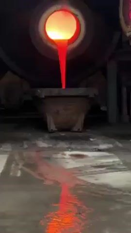 A furnace pours out molten metal into a crucible in a dark factory setting. The metal glows bright orange and red and forms a molten stream that continues to spill onto the factory floor. The floor appears wet and reflects the intense light of the flowing metal. The surrounding environment is dark and industrial, with large metal machinery visible.