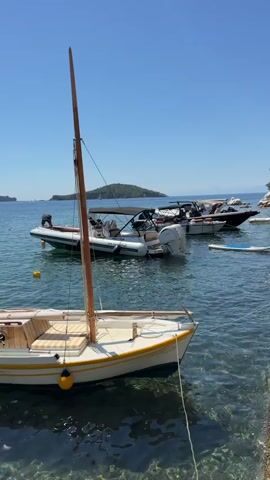 The video shows a waterfront scene with several boats docked or anchored in clear blue water. The first boat in the foreground is a small, white and yellow sailboat with a wooden mast. Behind it, a larger inflatable boat is visible with a person on board. Further out in the bay, other motorboats and a sailboat are present, with some appearing to be docked at a marina. The background consists of a coastline with green trees and some buildings. The sky is clear and blue, indicating a sunny day. The water is calm and transparent, allowing the seabed to be seen in some areas.