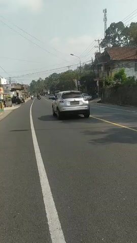 The video shows a view from a vehicle traveling on a paved road. A white SUV is in front, traveling in the same direction. There are other vehicles on the road, including motorcycles and cars. The road is lined with trees and buildings. The sky is clear and the sun is shining. The video is filmed from a low angle, suggesting it may have been taken by someone on a motorcycle or bicycle.