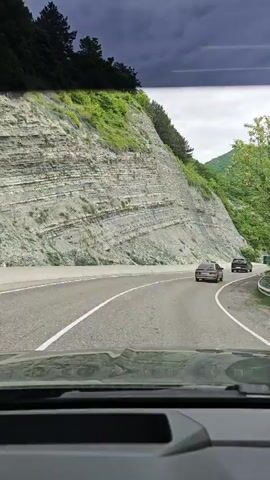 The video shows a car's point of view while driving on a highway. The road is asphalt with white lines marking the lanes. To the left of the road is a tall, layered rock face covered with some greenery. On the right are lush green hills. There are a couple of cars visible on the road ahead. A billboard is visible on the side of the road with some text in Russian. The sky is cloudy. The overall environment suggests a mountainous or hilly region. There is music playing in the background.