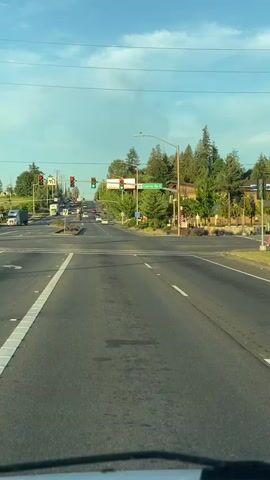 The video shows a view from a vehicle driving on a road towards an intersection. The road is paved and has white lane markings. Ahead, there is a traffic light and a street sign indicating "Garrision Rd". Several cars are stopped at the intersection. Trees and buildings are visible on the sides of the road. The sky is blue with some clouds.