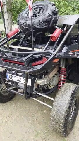 A black Polaris RZR XP 1000 side-by-side all-terrain vehicle is shown from the rear passenger side, parked on a dirt path. The vehicle has a spare tire mounted on the rear rack. The side of the vehicle displays the Polaris logo and "RZR XP 1000". Red suspension springs are visible on the rear. The environment appears to be a wooded area with some wooden structures in the background. Other ATVs are parked in the distance. The ground is muddy.