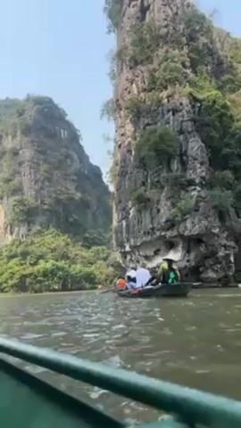The video captures a boat journey through a narrow waterway bordered by towering limestone cliffs covered with lush green vegetation. The perspective is from a boat, looking ahead towards another boat carrying several people. Some of the passengers are wearing life vests. The overall environment suggests a serene and scenic natural landscape. The weather appears to be clear and bright.