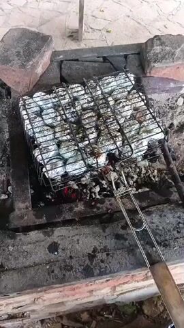The video shows a grill basket filled with food wrapped in aluminum foil, cooking over hot coals. The basket is placed in a brick pit. There are red embers visible beneath the food, indicating heat. The food appears to be arranged in neat rows within the basket. A metal handle extends from the basket, suggesting it is being used for cooking or transport. The background shows a stone or tiled surface and some rough-hewn bricks. The lighting suggests it is daytime, possibly outdoors.