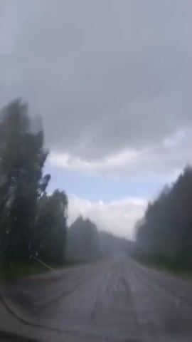 A first-person perspective video shows a dirt road covered in puddles, seen through a wet windshield. The road is lined with trees on both sides, and the sky is overcast with some patches of blue. The car appears to be moving, as the trees and road are blurred, suggesting motion. The overall atmosphere is one of a gloomy, rainy day.