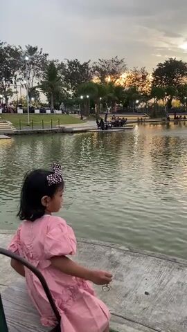 A young girl sits in a chair next to a body of water as dusk approaches. The girl wears a pink dress, a bow in her hair, and sandals. She looks at the water and turns to the camera, smiling. The body of water is calm and reflects the sky and surrounding light posts. Trees and people are visible in the background.