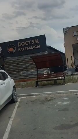 The video shows several cars parked in front of a building with signs. The first sign visible says "ДОСТУК КАТТАМАСЫ". A white SUV is parked in the foreground. A blue SUV is parked behind it, with a license plate visible. Another white SUV is parked in front of a building with a sign that says "НАМАЗКАНА" and "ЫСЫК КАТТАМА САМОРУЧНЫ ЧАЙЫ". A white van is parked to the right of the screen, partially obscuring another sign that says "Дария". There are people visible inside the buildings and standing near the vehicles. The environment appears to be an outdoor parking lot.