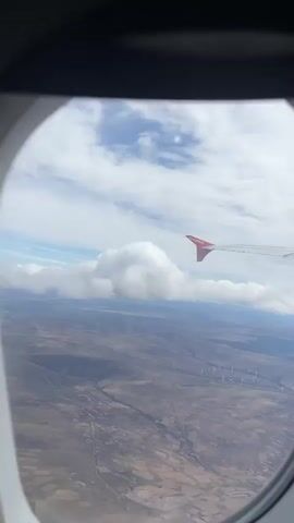 The view from an airplane window reveals a landscape below, with an EasyJet plane wing visible in the upper right of the frame. The land is mostly brown and gray, possibly an arid or rural area, crisscrossed with what appears to be roads and small winding streams. A wind farm of many wind turbines can be seen in the distance. The sky is a mix of blue and white, with fluffy white clouds scattered across the horizon. The camera is stationary, showing the ground receding as the plane continues to move.