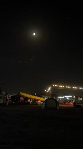 A nighttime scene at what appears to be a beach or outdoor gathering area. A full moon is visible in the dark sky. In the foreground, there is a dark, sandy or gravelly ground with a small tent set up. In the middle ground, there are several objects that appear to be recreational equipment, possibly kayaks or rafts, stacked together and covered with a yellow tarp. Behind this, a string of lights illuminates a low structure or seating area where several people are visible, sitting or reclining. The overall atmosphere is dim and nocturnal, with artificial lighting from the string lights providing contrast against the darkness.
