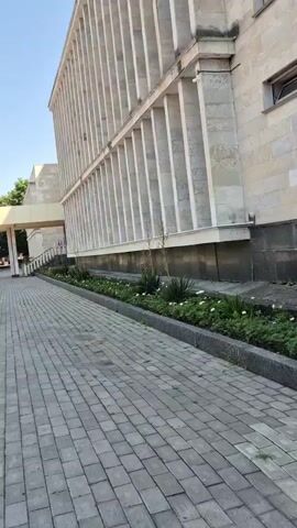 A low-angle shot pans across a gray paved walkway in front of a large, modern building. The building's facade is made of light-colored stone and features a series of vertical white columns casting shadows. Beneath the columns, a narrow garden bed runs along the building's base, filled with green foliage and white flowers. The walkway is made of square gray pavers, some of which have small weeds growing between them. The sky is a clear blue. In the distance, a covered walkway leads to an entrance of the building. The scene is quiet and still, with no people visible in the immediate frame.