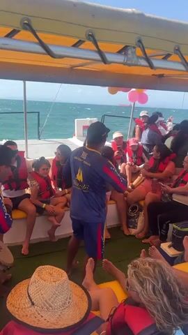 The video shows a group of people on a boat, presumably a catamaran, on a sunny day. Most of the passengers are seated on cushioned benches, many wearing life vests. There are balloons visible near the front of the boat. Two men in matching blue shirts with a Venezuelan flag motif and the words "Catamaran Mamale" are standing. One man is facing the group, appearing to speak or entertain, while the other is standing to the side, listening. The water is visible in the background, and the overall atmosphere seems relaxed and celebratory.