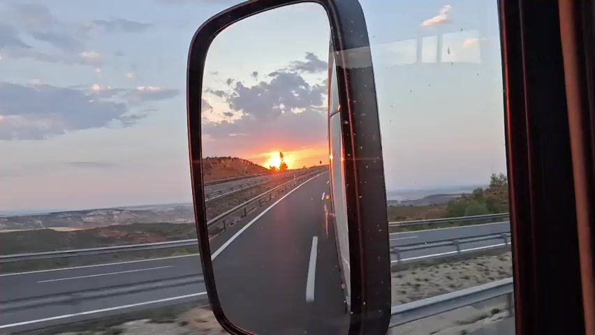 This video shows a sunset as viewed from the side mirror of a vehicle. The mirror reflects a highway with multiple lanes and a barrier on the side. The sun is setting behind a hilly landscape with trees. The sky is filled with clouds and the colors of the sunset are orange and pink. The scene appears to be a rural or semi-rural area. The reflection in the mirror is clear and detailed.