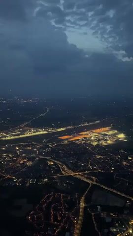 The video shows an aerial view of a city at night. The city is illuminated by a network of lights, including streetlights, building lights, and what appears to be an airport with bright runway lights and illuminated buildings. The sky above is dark with scattered clouds. The perspective suggests the camera is either from a plane or a drone, looking down on the sprawling urban landscape. Roads and highways are visible, distinguished by lines of headlights and taillights. Residential areas with smaller clusters of lights are also discernible. The overall scene depicts a busy, illuminated city under a twilight or nighttime sky.