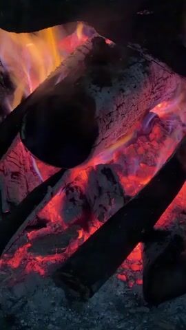The video shows a close-up of a campfire burning. Several dark logs are present, surrounded by glowing embers. Flames are visible at the top of the frame, showcasing shades of yellow, orange, and purple. Smoke is rising from the burning wood. The overall environment appears dark. A voice is heard speaking in the background.