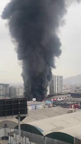 The media file captures a dramatic scene of a large fire erupting in Breña, Peru. A massive plume of thick, black smoke billows high into the sky, dominating the frame. The smoke originates from a building or group of buildings in the mid-ground, with visible flames appearing to emerge from within the structure at its base.

The setting appears to be an urban environment with a mix of industrial or commercial buildings and taller residential or office buildings in the background. In the foreground, a large billboard structure is visible, partially obscuring the view of some of the lower buildings. The weather seems overcast, with a hazy sky that could indicate either cloudy conditions or the effects of the smoke itself. The time of day is difficult to ascertain precisely due to the overcast conditions, but it appears to be daytime.

No people are clearly discernible within the immediate vicinity of the fire's origin. The overall emotion conveyed by the image is one of alarm and significant disruption due to the intensity of the fire. There is no visible text that provides specific information about the event. The scene is characteristic of a major industrial or structural fire within a populated area.