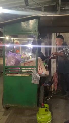 A man wearing a blue and white striped shirt, dark pants, and a black skullcap is standing by a green food cart. He holds a dark-colored tool, possibly a spatula or tongs, and appears to be interacting with food inside the cart. He is looking towards the cart and seems to be in the process of cooking or preparing food. To his right, another man in a white t-shirt and a grey baseball cap is visible from the back, facing away from the camera and looking towards the food cart. In the foreground, a green propane tank is visible on the ground. The environment appears to be an outdoor food stall, possibly at night, with artificial lighting illuminating the scene. There are plastic bags and other items stored around the cart. The overall impression is of a busy food vendor at work.