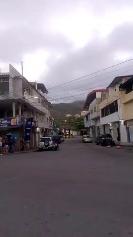 The video captures a street scene under a cloudy sky. Buildings line both sides of the paved road, some with shops on the ground floor. Several vehicles are parked or moving on the street, including cars, a motorcycle, and a sport utility vehicle. In the background, a hillside with vegetation and some buildings is visible. Power lines are strung across the sky above the street.