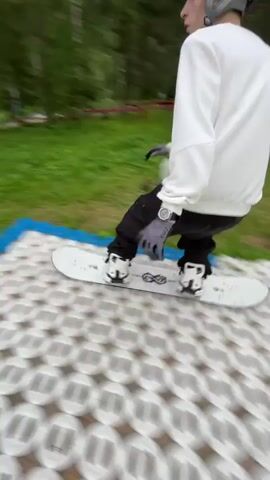A person wearing a white hoodie and black pants is snowboarding down a grassy slope. The snowboarder is in mid-air, performing a trick over a large black pipe laid on the ground. The pipe is partially covered by black mats, and there is a blue ramp leading up to it. The background shows a lush green landscape with trees and houses. The sky is cloudy. The footage is shaky and appears to be captured from a first-person perspective. The snowboarder lands successfully after the jump.