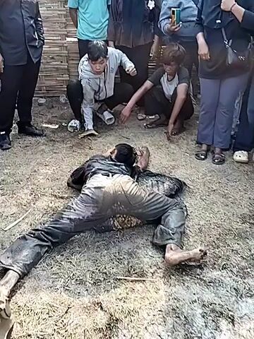 The video shows a man lying on his back on a patch of dry grass and dirt, with his arms and legs spread out. The man is wearing dirty gray pants and what appears to be a dark shirt or jacket. His feet are bare. 

Behind and surrounding the man, a group of about ten people are standing or crouching. Two people are crouching close to the prone man, appearing to touch or examine him. Other people in the group are standing and watching, some holding phones and possibly recording.  A makeshift bamboo fence is visible in the background. The overall atmosphere suggests a potentially serious incident or accident.