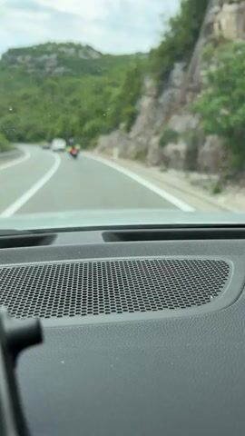 The video shows a view from inside a car, looking out the windshield while driving on a highway. The foreground is dominated by a textured black dashboard with a grille-like speaker cover. The road stretches ahead with painted white lines separating lanes. On the left, cars are moving in the same direction, and a motorcycle can be seen in the distance. To the right of the road, a metal guardrail runs along the edge, and beyond that, there's a rocky hillside covered in green vegetation. A mountain covered in trees dominates the background under a cloudy sky.