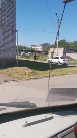 A person is seen walking towards a white car parked on a paved road. The person is dressed in dark clothing and is walking away from a brick building. The car is facing away from the viewer, and the driver's side door is open. A tall metal pole with wires is visible in the foreground, and a sticker is attached to the windshield. The background shows more buildings and trees under a clear blue sky.