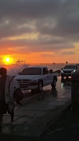 A white pickup truck drives on a wet road alongside the ocean at sunset. The sun is low in the sky, casting an orange glow on the clouds. Waves crash against the shore, and a pier stretches across the horizon. Another car is visible behind the pickup truck. A person is partially visible in the foreground on the left.