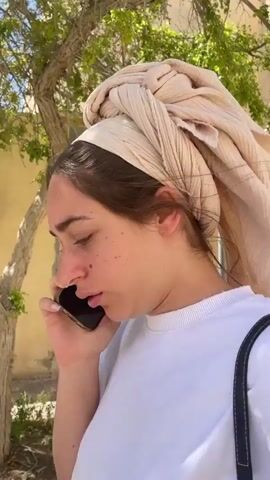 Here is a detailed description of the video:
A woman is standing outside, talking on a cell phone. She is wearing a white t-shirt and a beige head covering. She has brown hair and freckles. She is holding a black cell phone to her ear with her left hand. A shoulder bag is slung over her shoulder. A tree is behind her, and a building is visible in the background. A car passes behind her. The sky is blue.