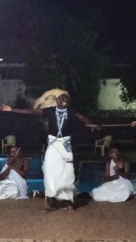 The video shows a nighttime cultural dance performance. A central figure, adorned in a black top, white wrap, a light-colored wig, and beaded necklaces, energetically leaps and dances in front of what appears to be a swimming pool. To each side of this dancer, seated and dressed in white fabrics, are two individuals who are clapping along to the rhythm. In the background, faint outlines of trees and buildings can be discerned under the dimly lit environment.