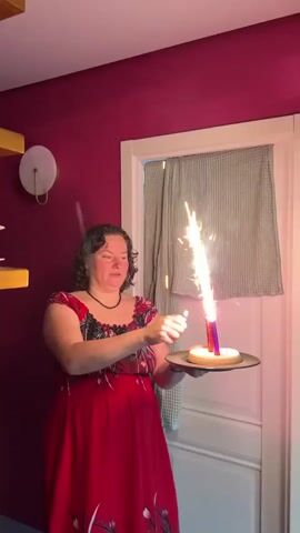A woman in a red dress holds a plate with a dessert and a lit sparkler candle. She is standing in front of a white door and a burgundy wall. The room has a wall lamp on the left side and a partially visible shelf with white objects. The woman seems focused on the sparkler candle, holding the plate with her left hand and gesturing with her right hand near the flame. The sparkler candle emits a bright fountain of sparks. The door has a neutral-colored dishcloth hanging on it. The overall lighting of the room is warm.