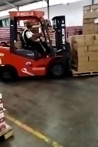 A red forklift with a driver is seen in a warehouse. The forklift is lifting a pallet of cardboard boxes. The warehouse has high ceilings and contains stacks of boxes.