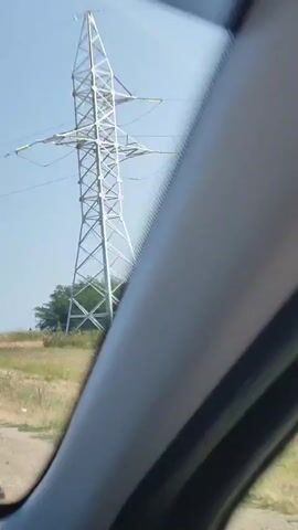 The video captures a view from inside a vehicle, looking out a window. In the foreground, a portion of the vehicle's interior trim is visible. The primary focus shifts to the exterior landscape, which features a tall, lattice-like electrical transmission tower. In the middle ground, there are green trees and vegetation. Further in the distance, a field with crops, possibly corn, is visible. The sky is clear and blue. The video also shows a wind turbine with rotating blades. The viewpoint appears to be moving, suggesting the vehicle is in motion.