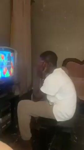 A man is seated in a room, facing a television set. He is wearing a white t-shirt and tan pants. He has his hands on his face, seemingly in distress or deep thought. The television displays a colorful animated scene. The room appears to be dimly lit, with drab walls and minimal decor. A curtain hangs to the side of the television. There is a small chair next to the man, and a stereo system below the television.